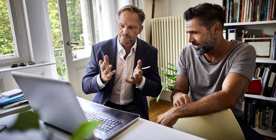 Two men seated at a desk, focused on a laptop, engaged in a discussion or collaboration.