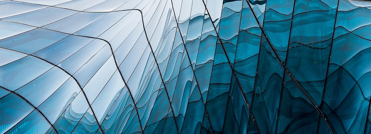 Close-up of a building facade with wavy, reflective blue glass panels, creating an abstract pattern with light and shadow