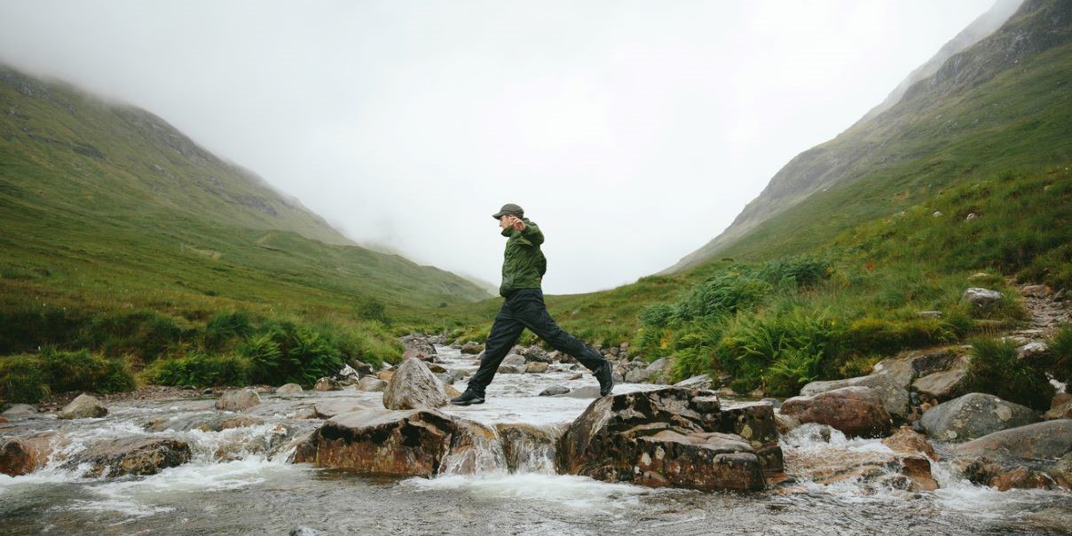 A man carefully crosses a mountain stream, surrounded by rugged terrain and lush greenery.