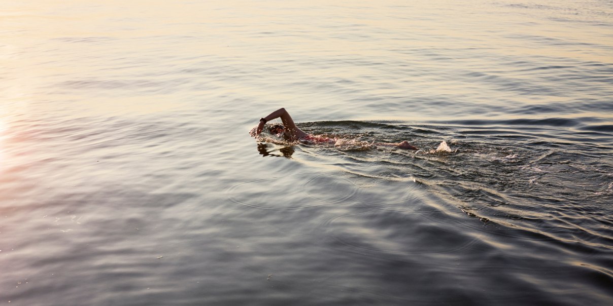 Swimmer performing freestyle stroke in calm water during sunrise or sunset.