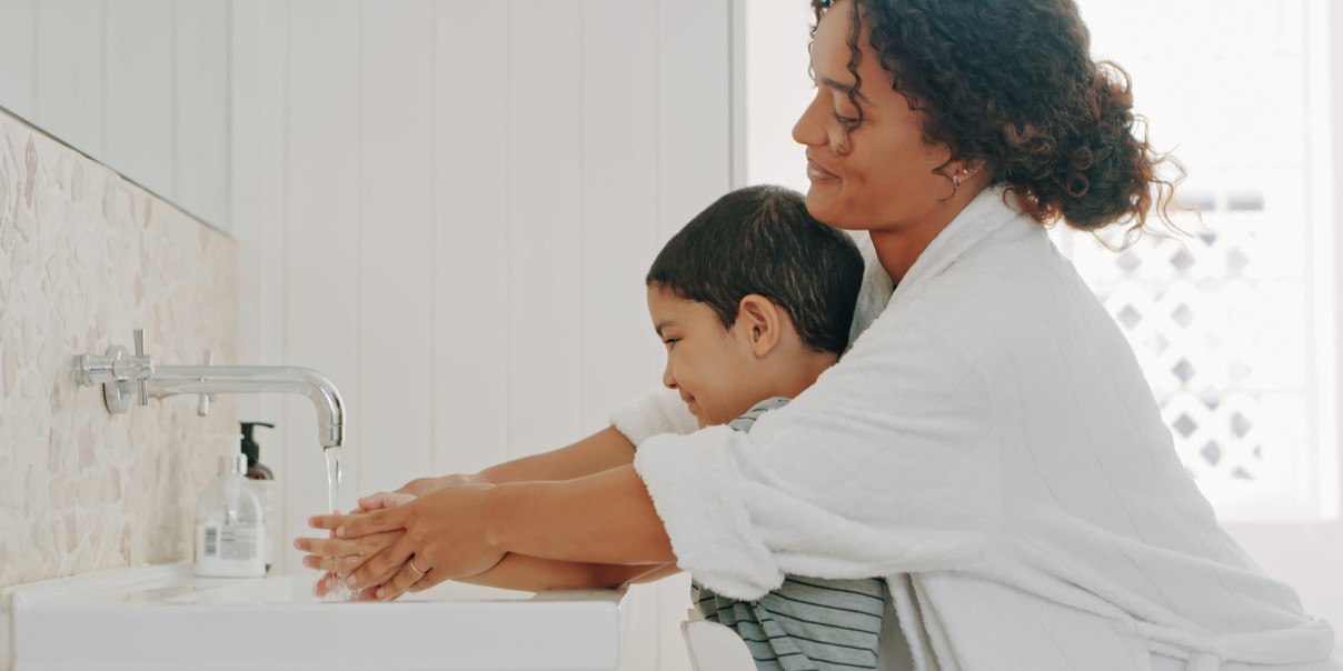 Adult helping child wash hands at a sink with running water and soap dispenser nearby.