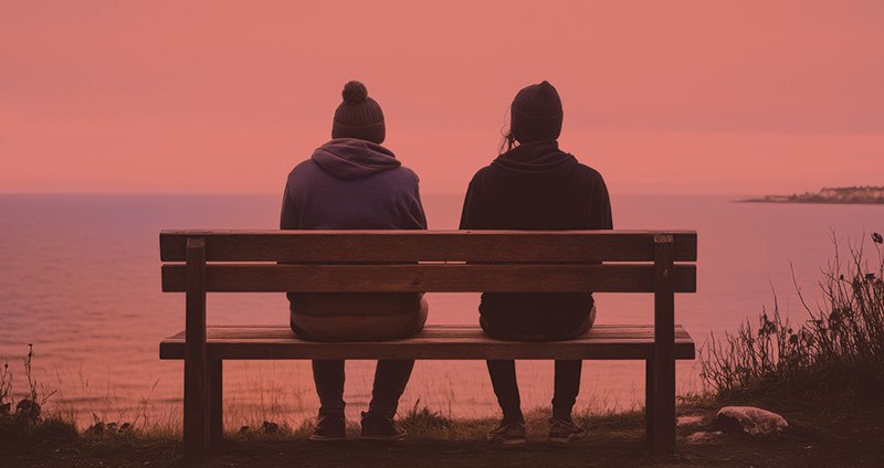 Two people sitting on a bench overlooking the ocean at sunset, with a reddish tint over the scene