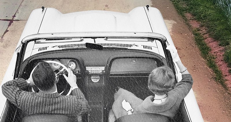 Top view of two people in a vintage convertible driving on a road beside grass.