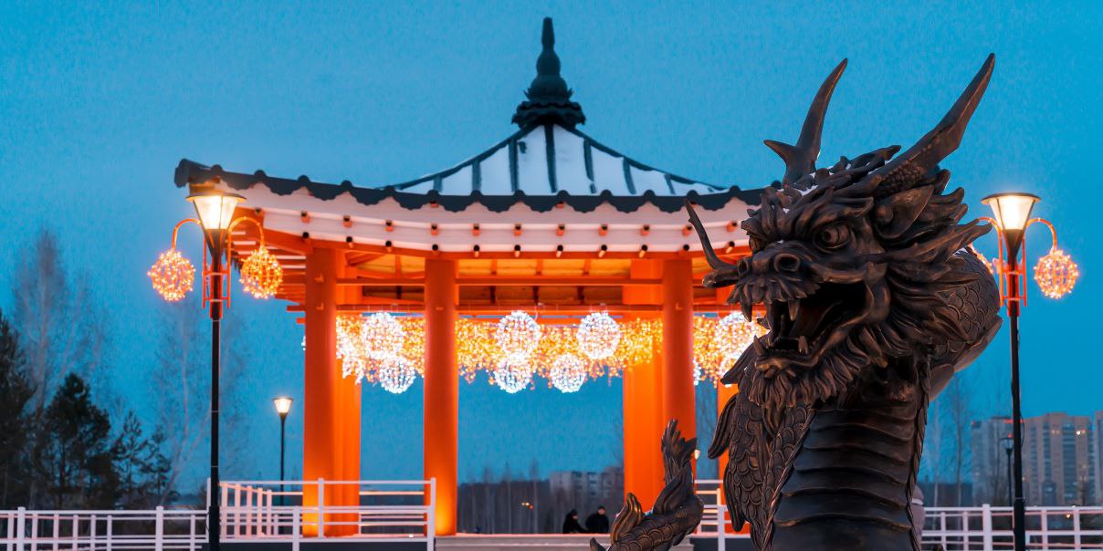 Traditional Asian-style pavilion with a black and white roof, orange columns, and hanging spherical lights, featuring a detailed dragon statue in the foreground against an evening sky.