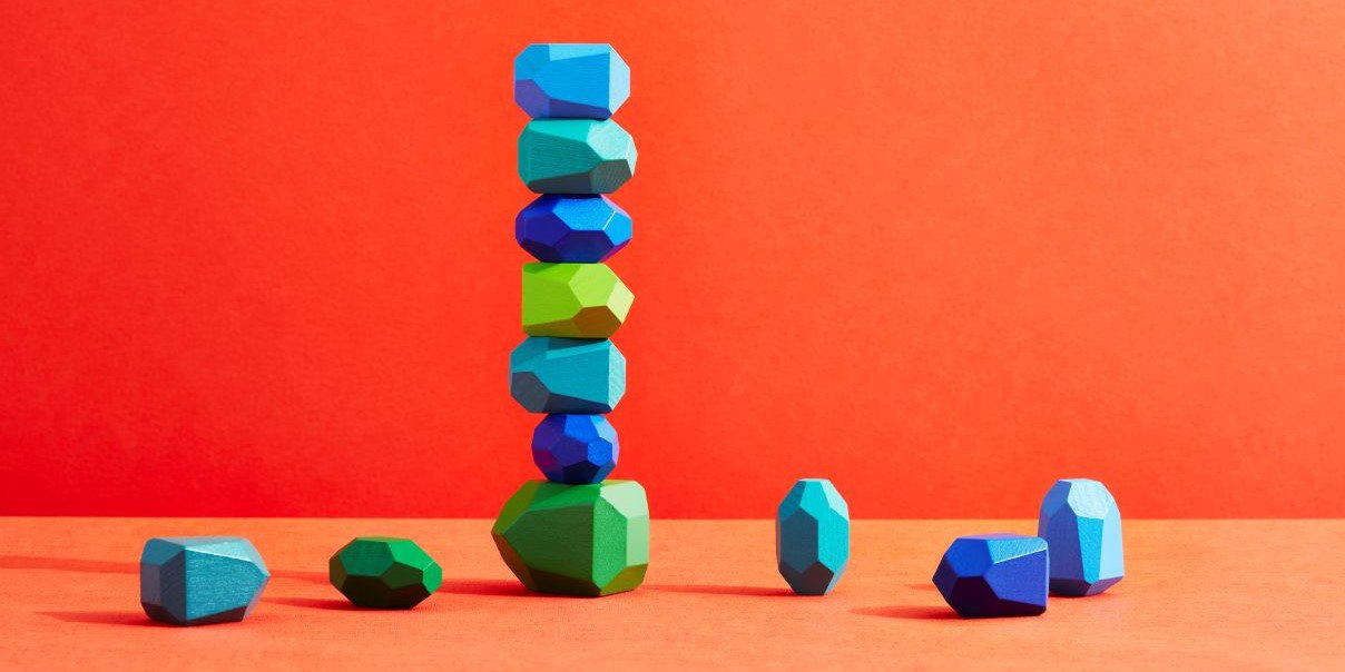 Colorful geometric stones stacked vertically on an orange background, forming a tall column with other stones scattered at the base.
