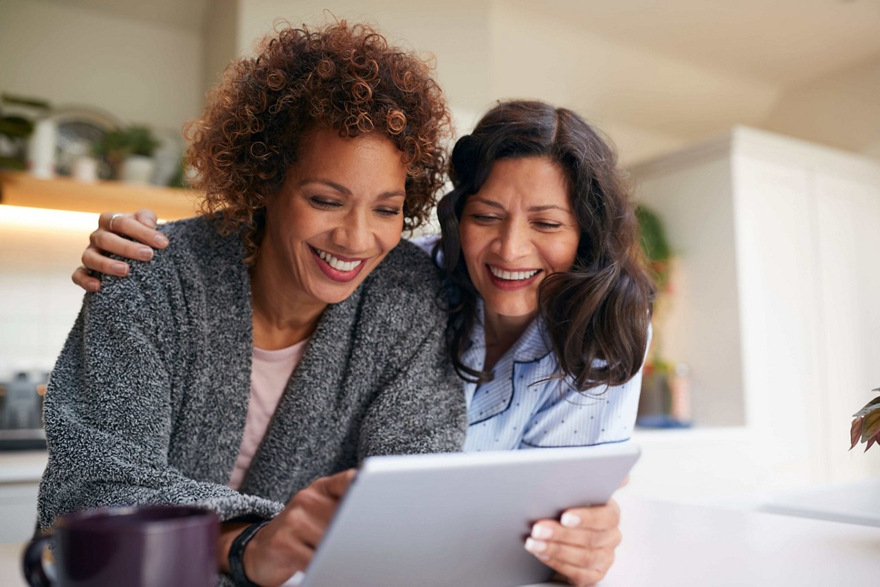 Mature Female Couple At Home In Kitchen In Pyjamas Drinking Coffee With Digital Tablet