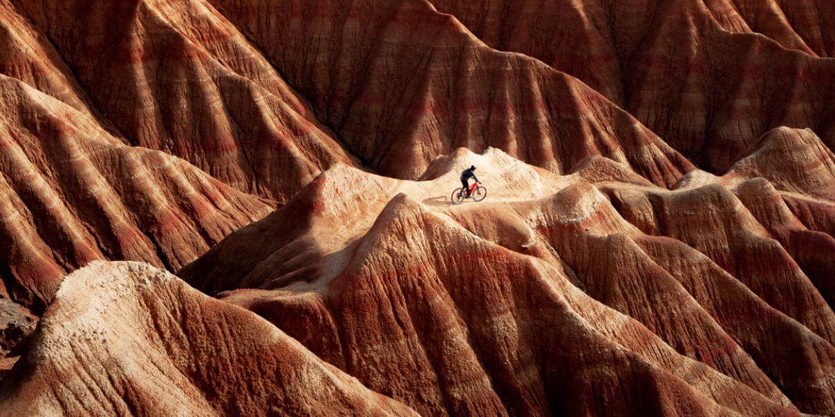 A cyclist riding through rugged, reddish-brown, mountainous terrain