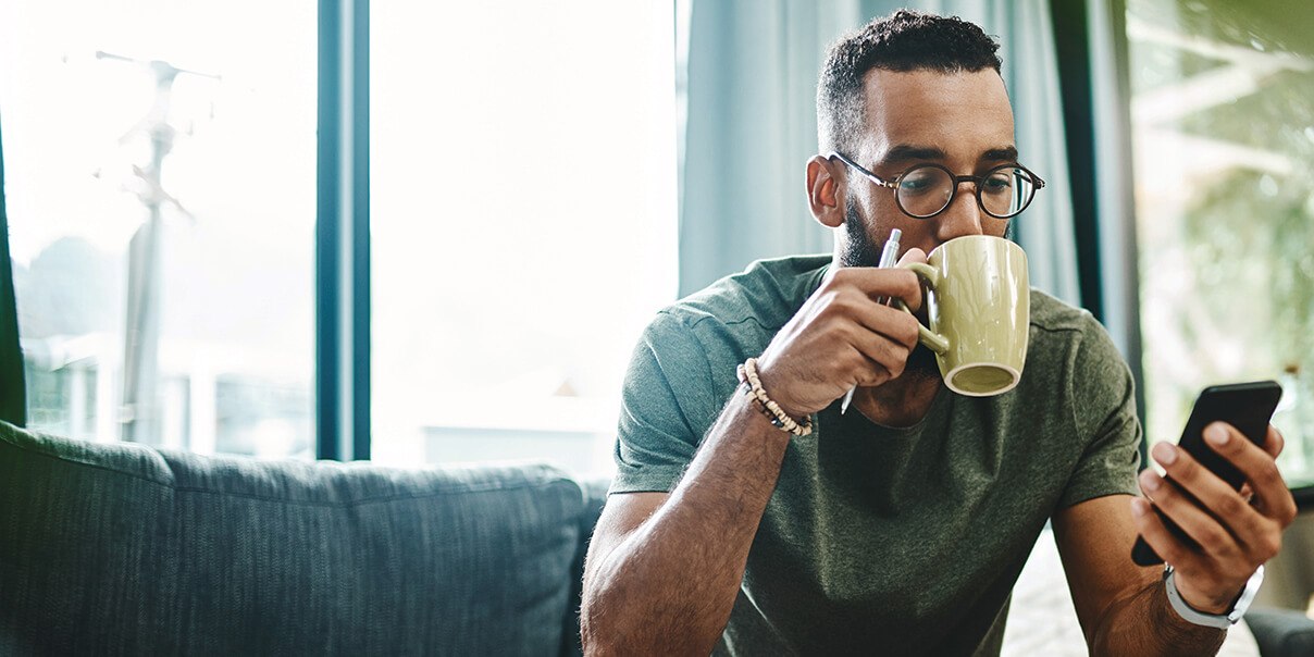 Shot of a young man using a smartphone and having coffee on the sofa at home