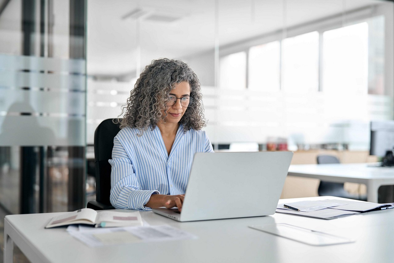 Serious busy mature middle aged professional business woman, older lady manager executive leader wearing glasses looking at laptop using computer in office working on digital project sitting at desk.