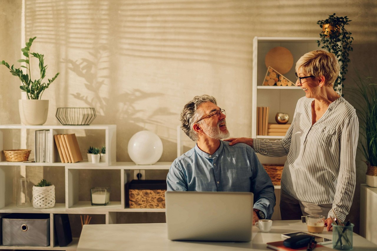 Portrait of a happy mature couple laughing and bonding while sitting at home at the table with laptop. Senior spouses having fun while browsing internet and spending their time together. Copy space.