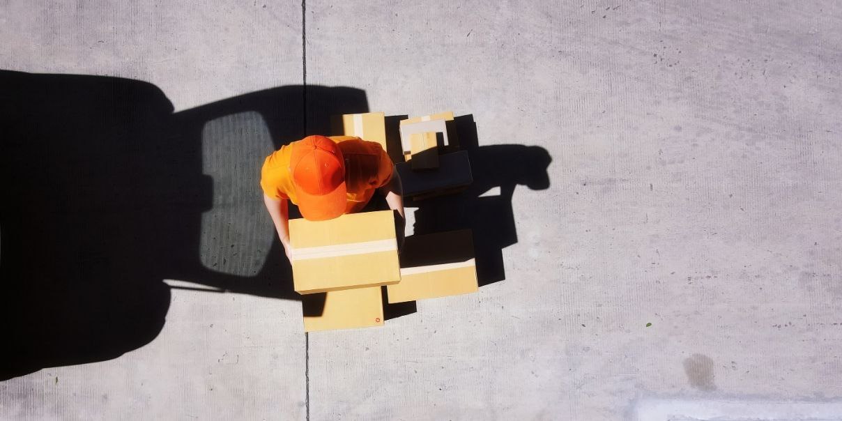 An overhead view of a person wearing an orange cap and shirt, handling cardboard boxes on a concrete surface. Shadows of the person and nearby objects are visible on the ground