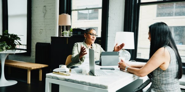 Two woman in an office setting. They are sitting at a desk as the one on the left is explaining details to the one on the right.