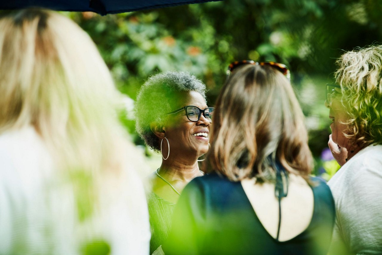 A group of women engaged in conversation at an outdoor event, surrounded by greenery and a lively atmosphere.