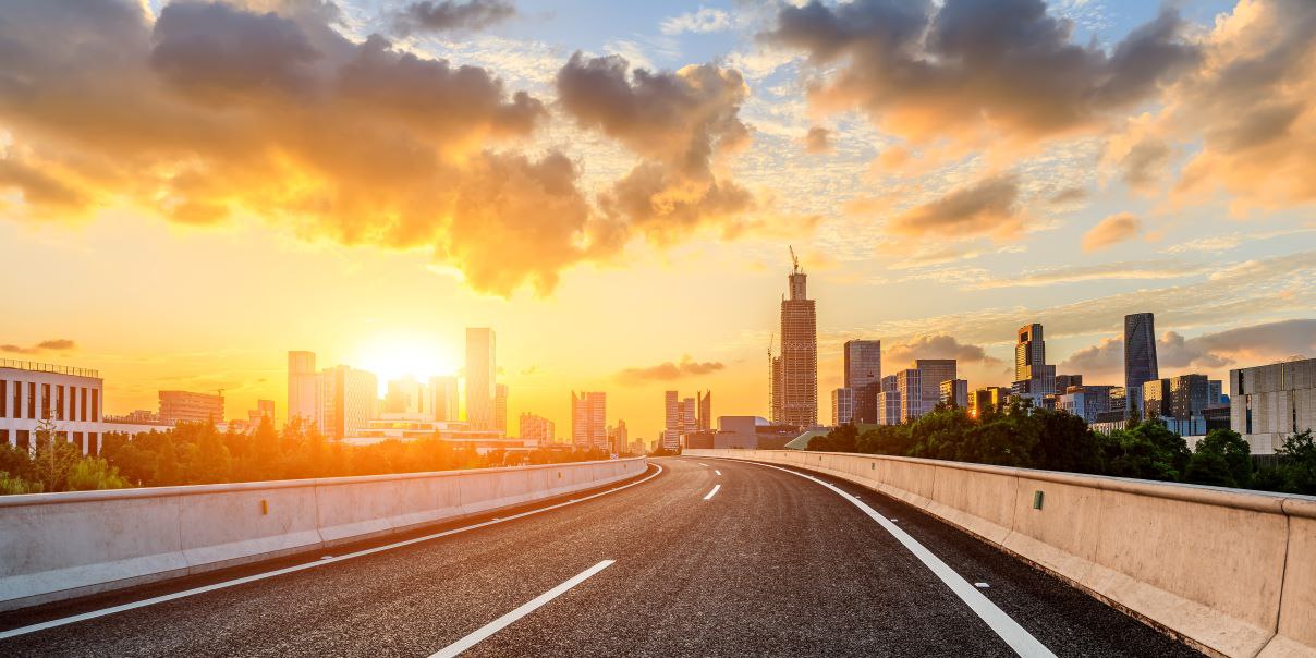 Modern cityscape at sunset with a curved road in the foreground, dramatic clouds, and tall skyscrapers illuminated by golden sunlight.