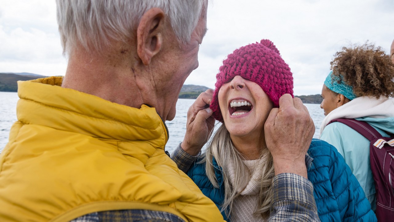 A mature couple enjoying a weekend away together in Torridon, Scotland, they are on a tourboat, enjoying an excursion out at sea. The man is pulling his wife's hat over her eyes while she laughs.