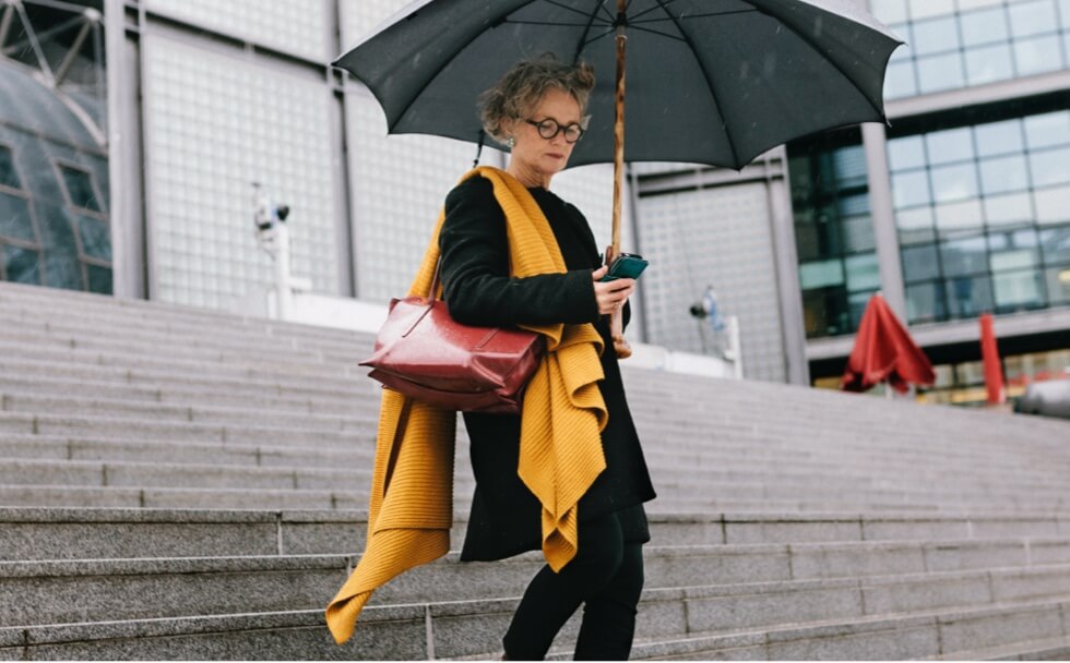 Woman holding a phone and a large umbrella walking down steps outside an office.
