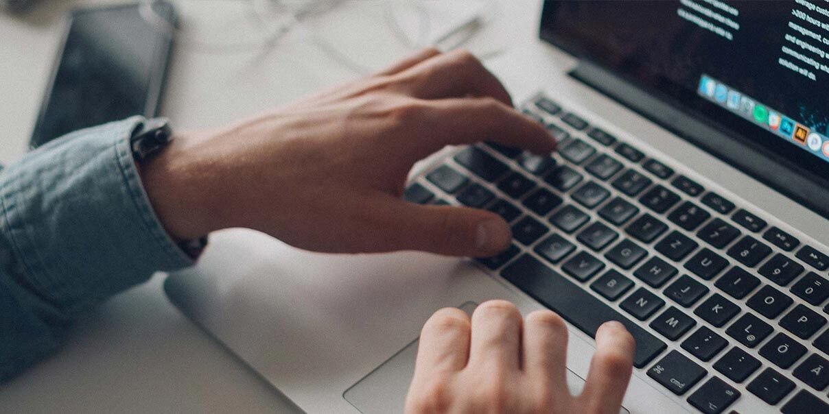 The hands of a person in a long sleeve shirt shown typing on a Macbook keyboard